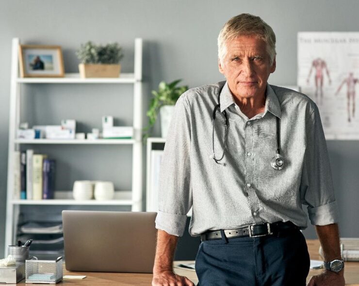 A doctor leans on his desk, looking forward