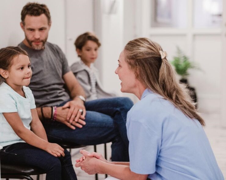 Female pediatrician talking with girl sitting by family in hospital corridor