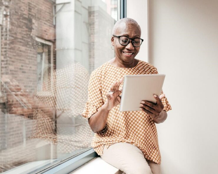 A senior woman sitting on a window-sill looking at a tablet