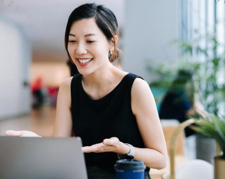 Smiling professional young Asian businesswoman talking in front of the camera having video conference with her business partners on laptop in a contemporary office space