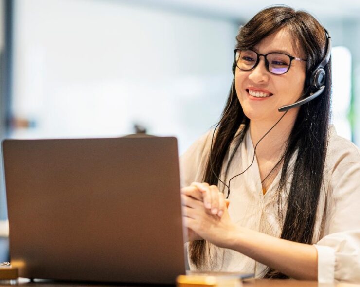 Asian woman with long black hair, sitting in front of an open laptop, wearing a headset and glasses, smiling.