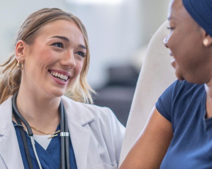 A female physician is wearing a white lab coat and holding out a clipboard as she talks to her patient. The patient is seated in a high-back chair and wearing a headscarf .