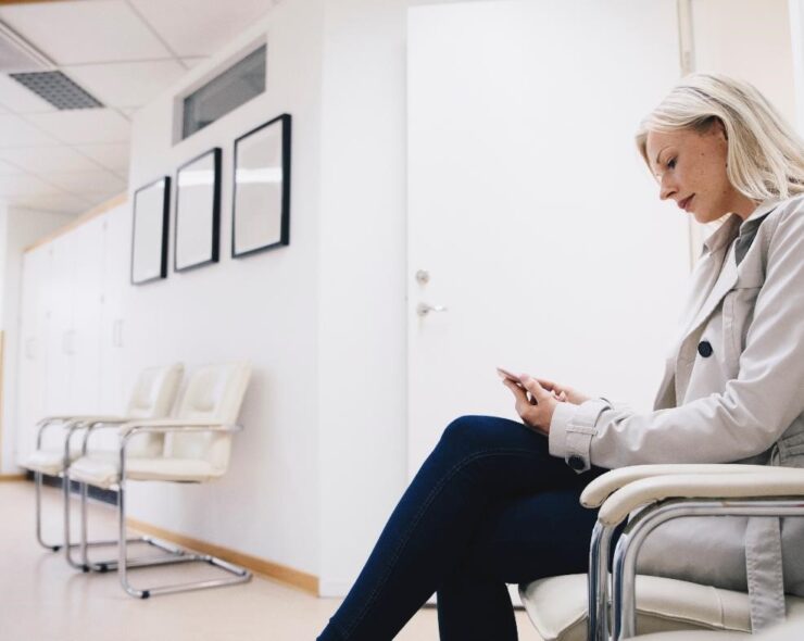 Side view of woman using smart phone while sitting on chair in hospital corridr